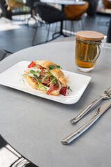 Croissant sandwich with prosciutto, berries, dijon mustard, cheese and cup of tea on white plate and bright background. Continental style breakfast.