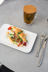 Croissant sandwich with prosciutto, berries, dijon mustard, cheese and cup of tea on white plate and bright background. Continental style breakfast.