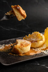 Hand with fork of Cottage cheese pancakes. Syrniki or Cheesecakes with dried pineapple and caramel isolated on black marble background. Homemade food. Tasty breakfast. Selective focus.