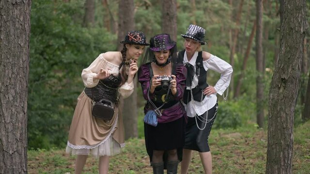 Zoom out three happy beautiful women in steampunk costumes watching photos on camera smiling talking in slow motion. Wide shot excited Caucasian ladies celebrating Halloween in forest