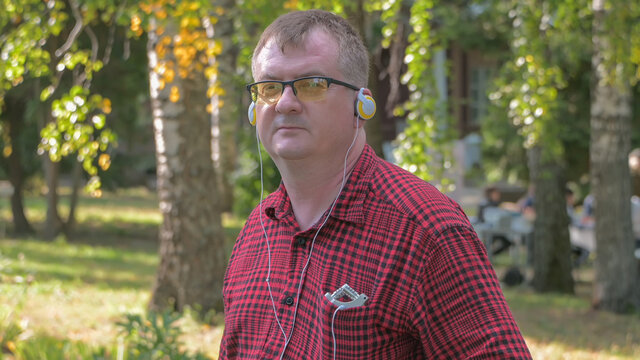 a man in a red shirt and glasses listens to music with headphones in nature in sunny weather against the background of autumn trees - Powered by Adobe