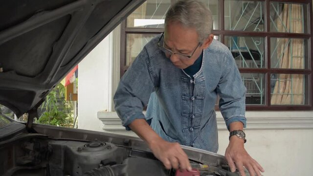 Asian elderly gray hair man in eyeglasses checking engine parts condition of an old car in front of his house. Retirement life.