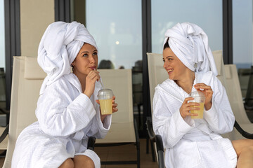 Two happy attractive brunette female friends in white bathrobes and white towel on head have fun on...