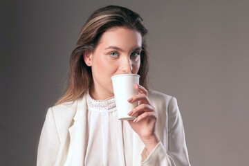Attractive girl holds a pure white glass - mockup for paste design, blank glass for drinks. Studio portrait of cute woman, model in white clothes looking straight. Blurred details, grey background.