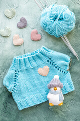 The concept of knitting clothes for toys and babies. A knitted T-shirt made of turquoise cotton yarn. A ball of yarn, knitting needles, toy and velvet hearts on a green background. Flat lay, vertical.