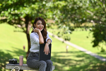 Photo of a beautiful woman holding a stylus pen and digital tablet while sitting and relaxing on the stone bench in the urban park.