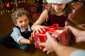 Fototapeta na wymiar Excited cute kids happily smiling, opening Christmas gifts. Beautifully decorated living room with lights and Christmas tree. Children having fun, celebrating family holiday