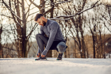 Sportsman crouching and tying shoelace in nature at snowy winter day. Sportswear, winter fitness, cold weather