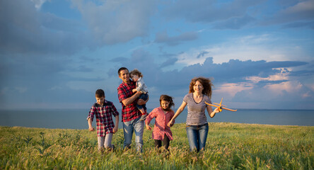 A large family with children launching a toy plane into the sky walking at sunset across a field with a blue sky background with clouds.