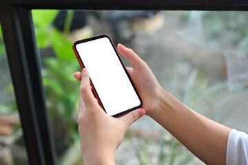 Cropped image of woman hand holding a white blank screen smartphone over the garden outside the windows as a background.