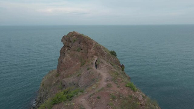 active tourist man with backpack walking along rocky mountain trail on sea coast shot from aerial view flying drone