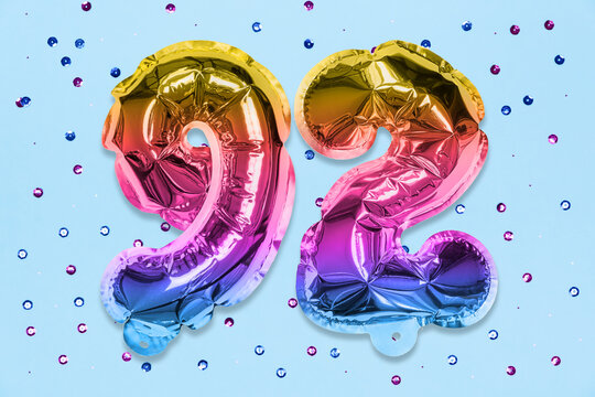 Rainbow foil balloon number, digit ninety two on a blue background with sequins. Birthday greeting card with inscription 92. Top view. Numerical digit. Celebration event, template.
