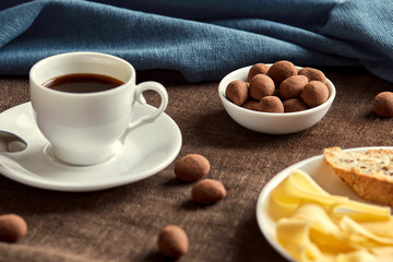 There are chocolates in a small plate. The plate is on a brown napkin. In front of the plate are a cup of coffee and a large plate of cheese and bread. Several chocolates scattered across the table.