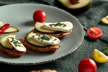 On the plate are toasts with avocado and feta cheese. Chopped vegetables and a tomato lie next to the toast. The plate is on a green tablecloth. Next to the plate are tomato slices and half an avocado