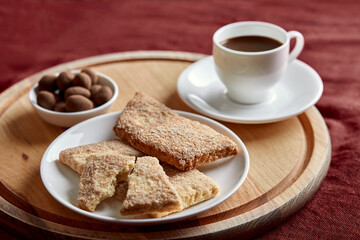There are cookies on the plate. Behind the plate are a small plate of sweets and a cup of coffee. The dishes are on the board. A wooden board lies on a red tablecloth.