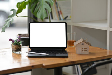 Photo of a white blank screen digital tablet with a keyboard case putting together with a stylus pen on a wooden working desk surrounded by a house model and small cup of plant.