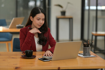 Portrait of a female business owner using a computer laptop at the wooden table surrounded by a coffee cup, smartphone, notebook and pencil holder.