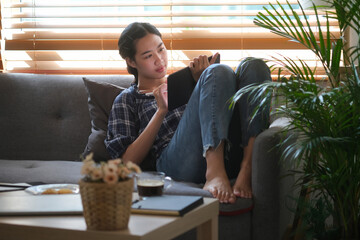 Photo of beautiful Asian woman using a digital tablet and stylus pen while laying on a cozy couch.