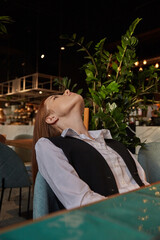 portrait of young caucasian woman with long hair in white blouse and black vest lying on chair at cafe with her chin up, throwing her head back. pretty lady posing at coffee shop