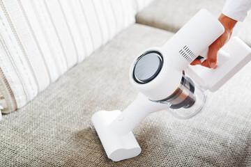 Fototapeta premium Powerful cordless vacuum cleaner with white cyclonic dust collection technology in hand, cleans the carpet in the house near the sofa.