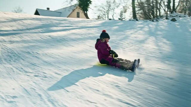 Tracking shot of a Caucasian little girl, with a warm knit hat, laughing descending the icy slope on the yellow shovel sledge.