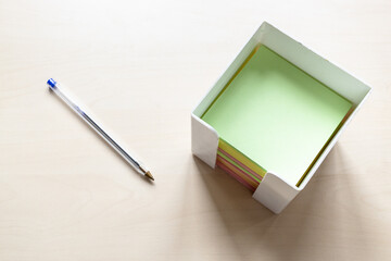 top view of pack of blank colorful sheets of note paper in box and simple blue pen on light brown wooden board