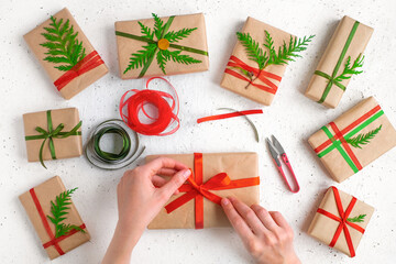 Fototapeta na wymiar Woman's hands wrapping christmas holiday handmade present in craft paper with red ribbon. Making bow at xmas gift box, decorated with fir branch. Scissors on white table, top view.