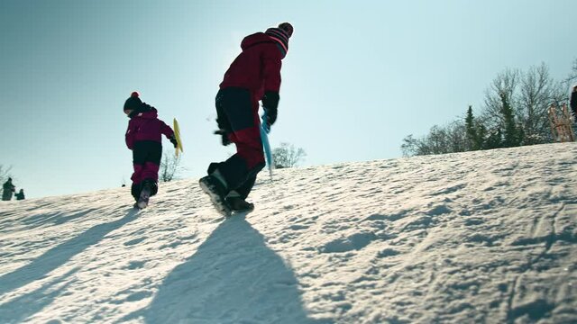 Handheld shot of a Caucasian family, father, son, and little daughter enjoying the winter sun, having fun on a sledding hill