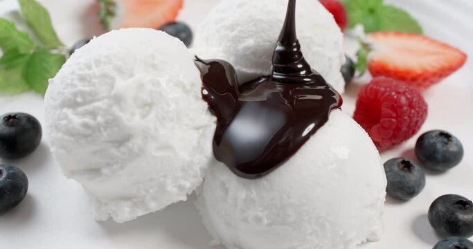 White vanilla ice cream scoops poured with chocolate sauce. Delicious ice cream and berries raspberries, strawberries and blueberries in bowl. Pouring topping