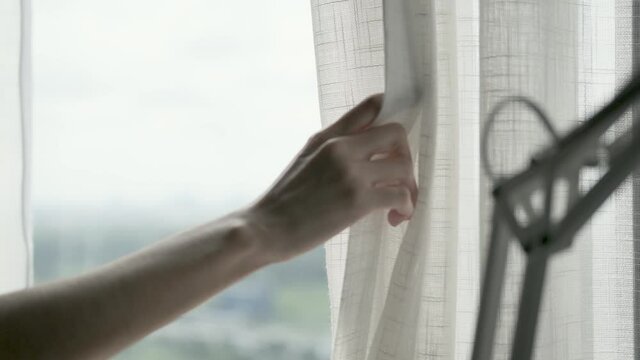 A woman's hand is opening the sheer curtains at the large window to let the light in during the day. 4k
