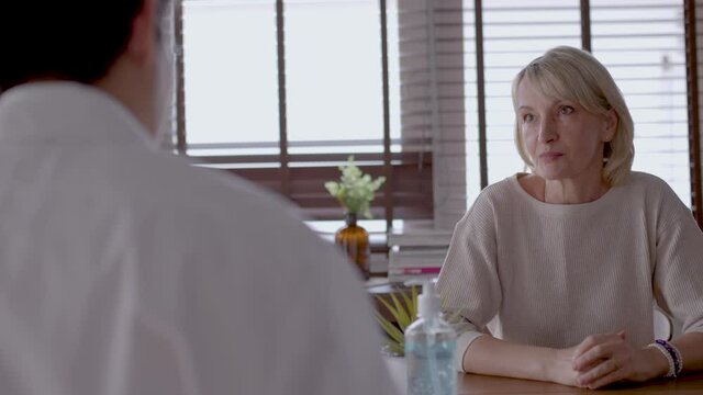 Senior caucasian woman consults with young asian doctor. Patient is describing about mental health problem to doctor. medium close-up with slide movement.  