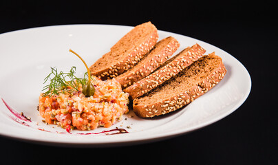 Salad with salmon and brown bread on a white plate on a black background. Tartar