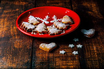 Christmas cookies in a red plate on a dark wooden table. Heart-shaped homemade baked goods sprinkled with white powdered sugar on top in a Christmas plate and signed 2022.