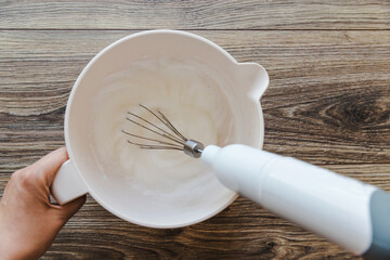 Beat egg whites with a mixer. A whisk blender whips the egg whites onto the tiramisu. A woman's hand holds a bowl of proteins for cream.
