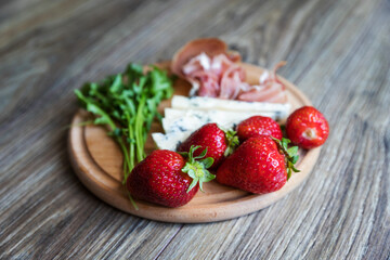 Dorblu cheese, jamon, arugula and strawberries on a round wooden board. Plateau with prosciutto and blue cheese, strawberries and fresh arugula on a wooden table background. Summer seasonal antipasto
