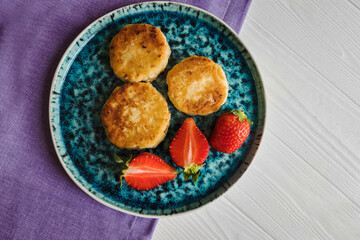 Сheese pancake, сheesecakes with fresh strawberries on a blue plate against the background of a lilac napkin and a white wooden table.
