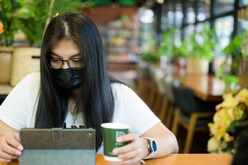 An adult Thai woman with a face mask is using digital tablet while sitting alone and drinking...