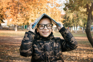 A boy with glasses in an autumn park holds a book on his head.