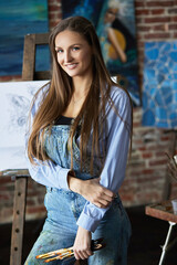 Portrait of young smiling female artist with her artworks in art studio. Painter holding art brushes. Creative process, relaxation, leisure, hobby, stress management.