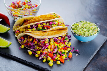 Vegetarian tacos with various vegetables, guacamole and sliced lime on dark background. Tacos with sweet corn, purple cabbage and pepper on a slate board. Copy space