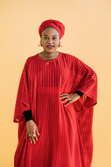 Women in traditional African style. Waist up studio shot of beautiful African woman in red dress, headscarf and jewelry posing against yellow background and looking at camera