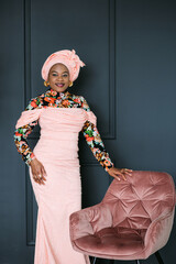 Gorgeous african woman in traditional dress with african print and headscarf, standing in studio near pink soft chair