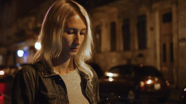 An attractive woman using a mobile phone While walking through the streets of a night city. The blonde is wearing a denim jacket. Slender thin girl.