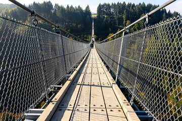 Suspension wooden bridge with steel ropes over a dense forest in West Germany, visible tourists on the bridge.