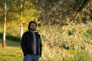 a young bearded man standing by the trees in the autumn park and looking thoughtfully at camera