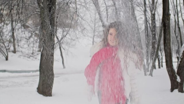 Happy young woman throwing up snow in winter park. Winter concept. Cute female in white jacket with red scarf enjoying beautiful winter day.