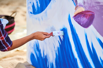 Female artist working on abstract acrylic painting, hand holding paint brush, large canvas outdoor