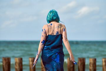 Artistic blue-haired woman performance artist smeared with blue gouache paints dancing on beach
