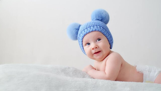 Happy newborn baby in a blue hat with pompoms looks into the camera. Smiling two-month-old infant is lying on a blanket. Beautiful, happy little child with blue eyes.  Cheerful White kid. SLOW MOTION