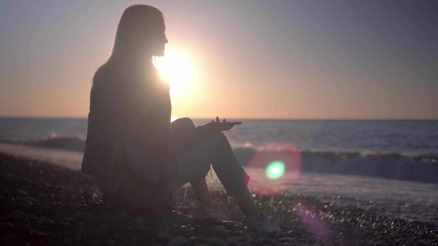 blonde in denim suit is sitting on seashore at sunset with a smartphone in her hands. She sits on rocks, communicates on social networks and shares her impressions. Talking to family and friends.